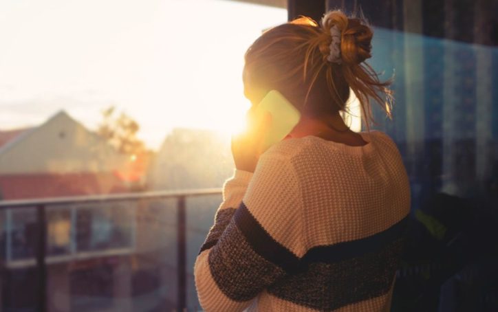 A person standing by a window talks on a smartphone while facing the sunset. The bright golden light silhouettes them against a view of distant buildings.