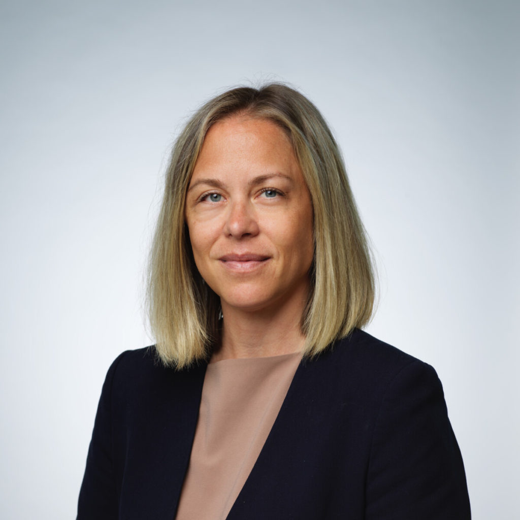 A portrait of a woman with shoulder-length blonde hair and a gentle smile. She wears a dark blue blazer over a tan top against a soft, light-gray studio background.